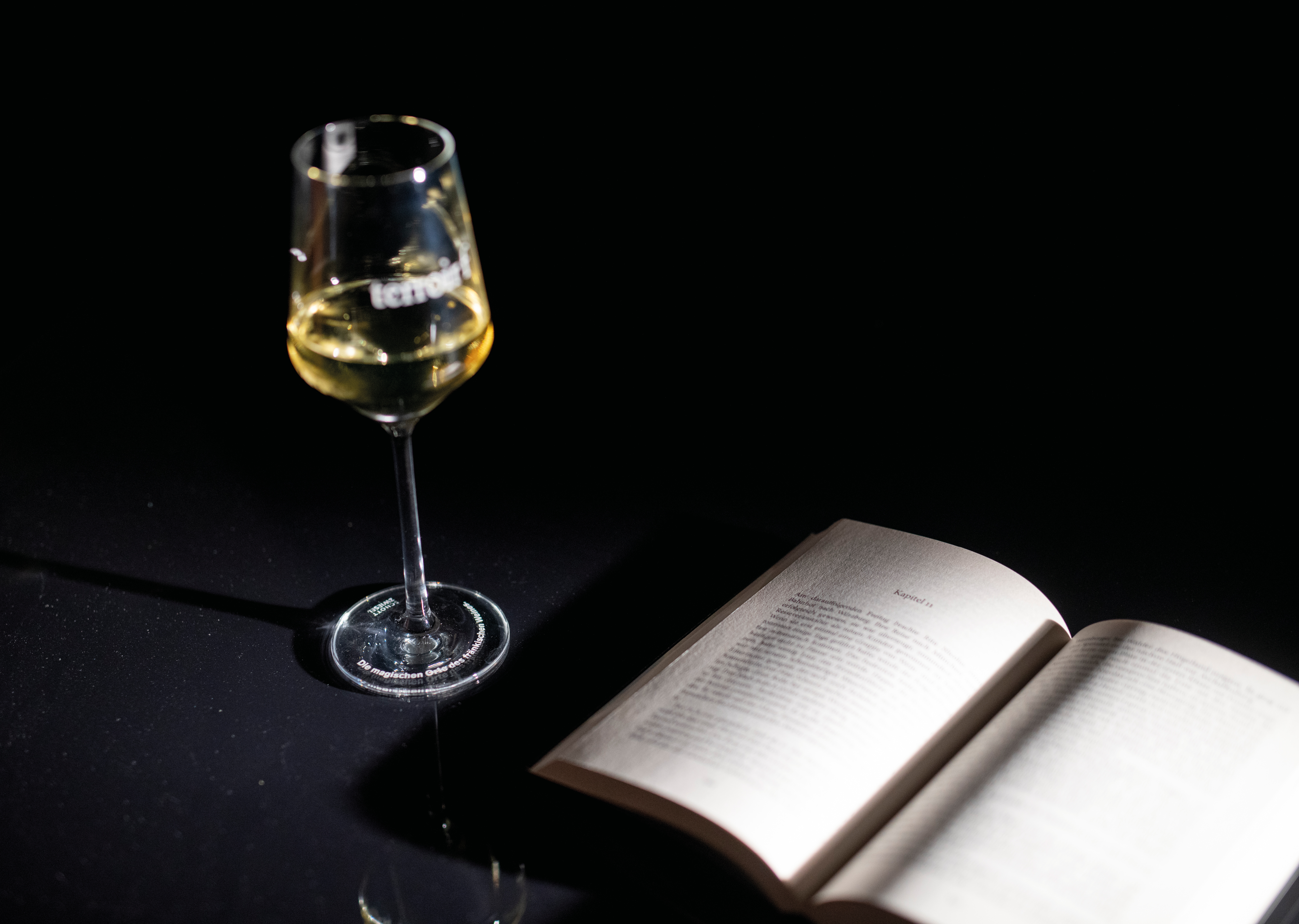 Ein halb gefülltes Glas Wein steht auf einem Tisch, daneben liegt ein aufgeschla-genes Buch.