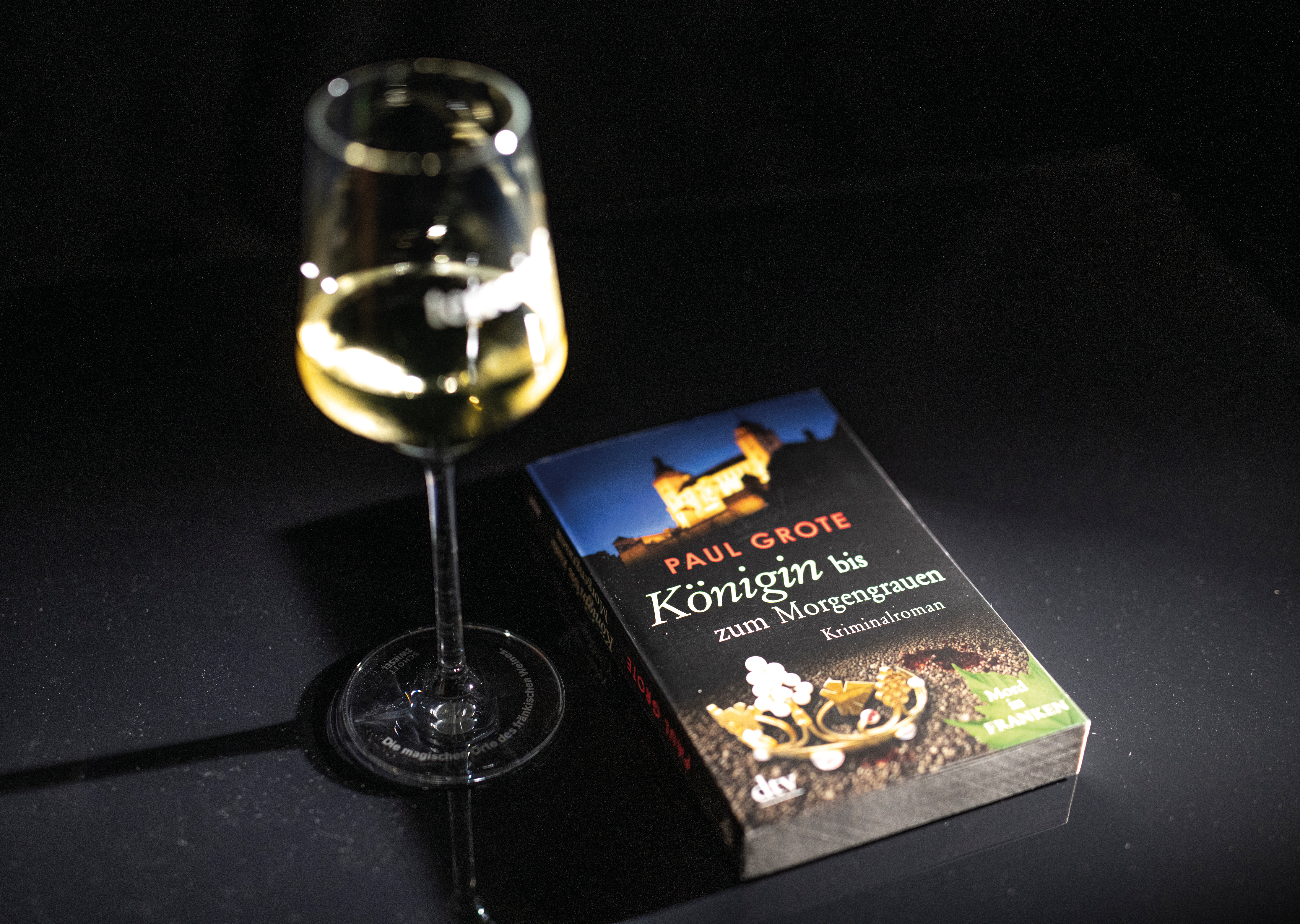 Ein halb gefülltes Glas Wein auf dem Tisch, daneben liegt ein Krimi.